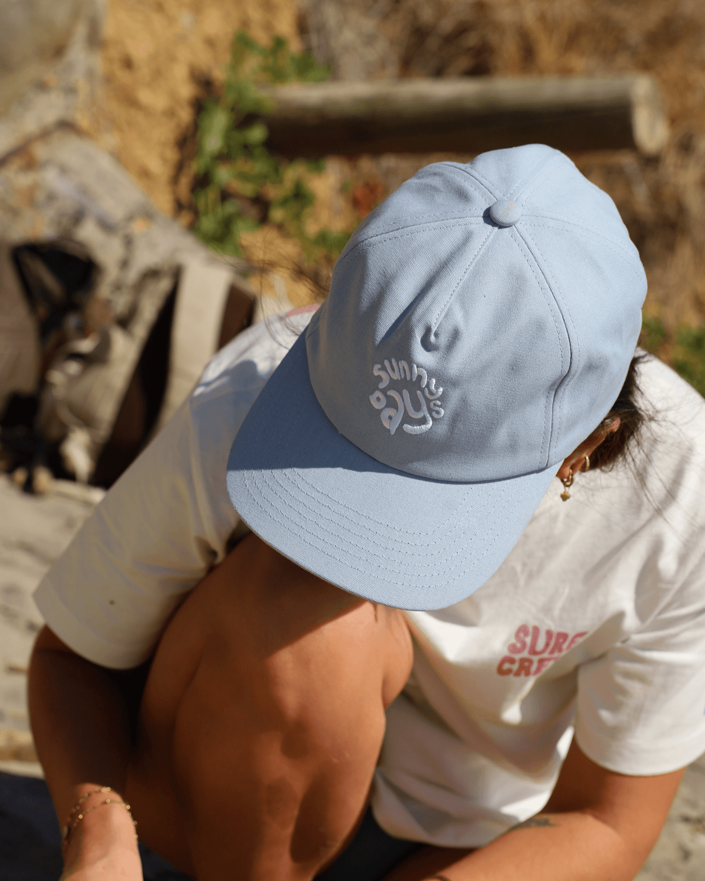 Casquette bleu pâle concu par lix pour les hommes et les femmes avec broderie sunny days au devant. Ajustable à l'arrière. Tshirt blanc avec imprimé surf crew rose au devant et gros imprimé surf crew rose et vague bleu au dos. 
