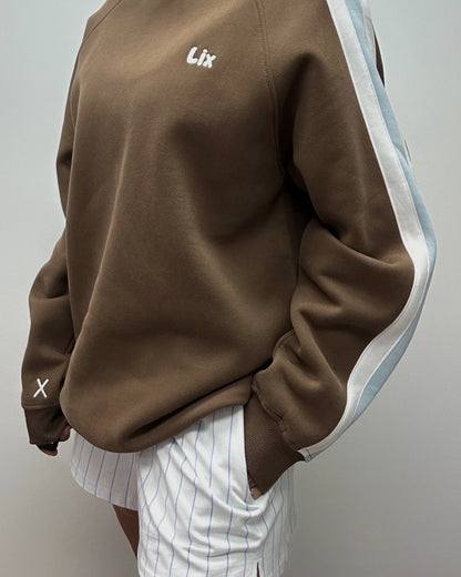 Crewneck brun avec broderie et rayure blanche et bleu sur les manches. Chandail brun avec manche de style raglan. 