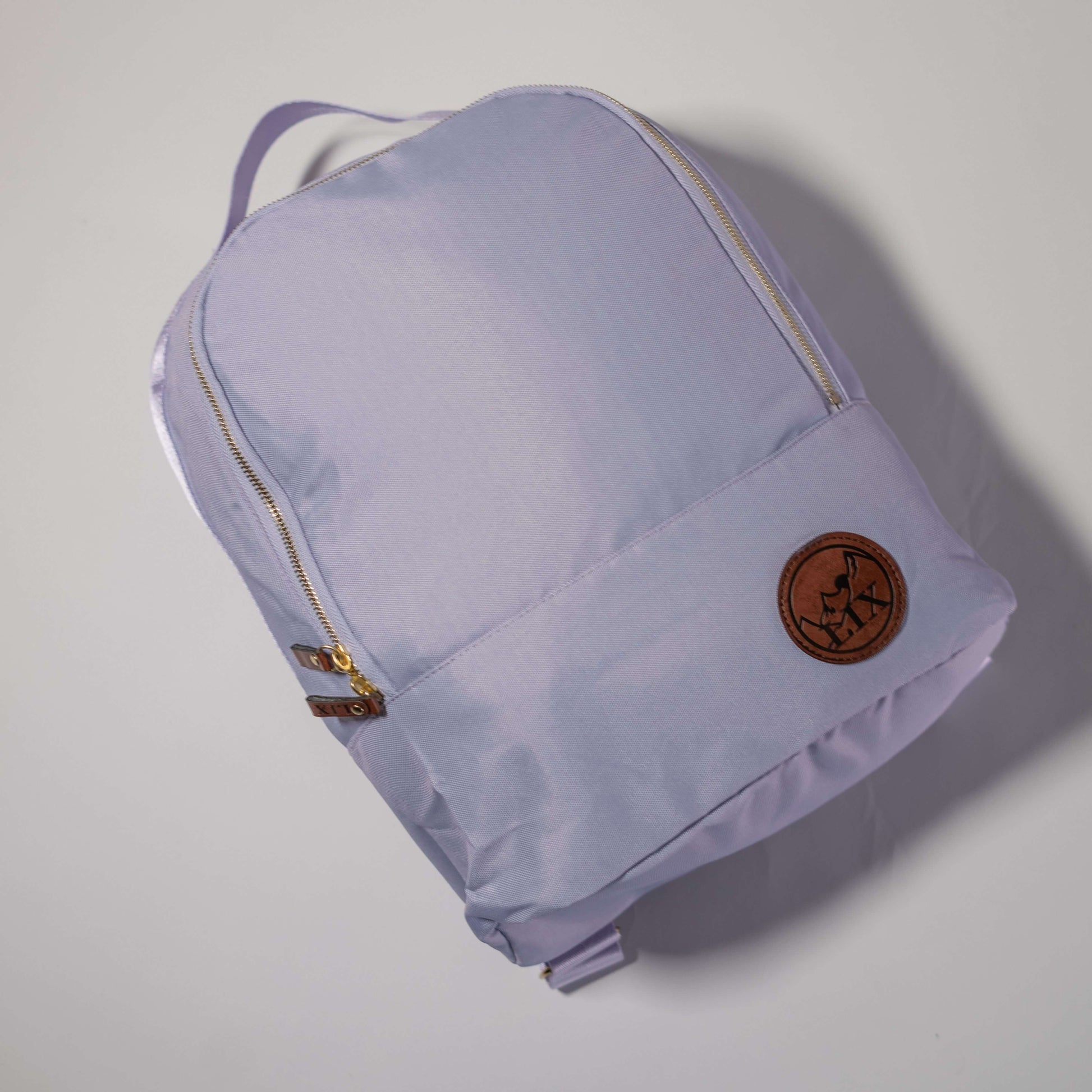 Sac à dos couleur lilas pale de conception éco-responsable. Badge en cuir synthétique végan de couleur brune avec le logo Lix design embossé en noir. Languette de fermeture éclaire en cuir végan brun avec Lix inscrit en noir.