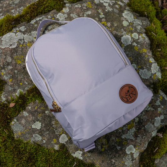 Sac à dos couleur lilas pale de conception éco-responsable. Badge en cuir synthétique végan de couleur brune avec le logo Lix design embossé en noir. Languette de fermeture éclaire en cuir végan brun avec Lix inscrit en noir.Backpack made in canada. Lix. Sac a dos Canada