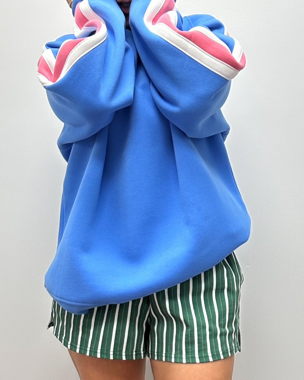Outfit look vintage année 90. Crewneck bleu avec rayure rose et blanche sur les manches. Short verte avec ligne blanche et brune. Look pour femme de 25-45 ans. 